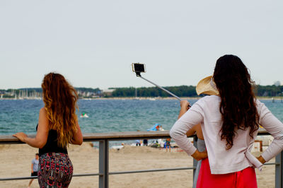 Rear view of friends taking selfie while standing by railing against sea