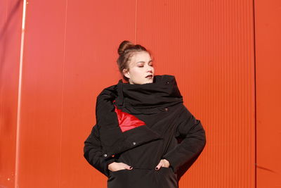 Fashionable teenage girl wearing black warm clothing standing against red wall during sunny day