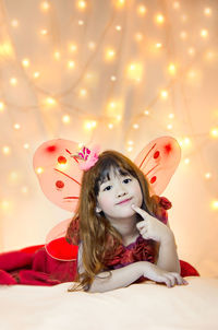 Portrait of smiling cute girl in angel costume lying on bed against illuminated lights