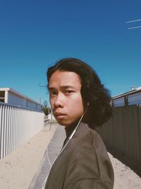 Portrait of serious young woman listening music while standing on road against blue sky