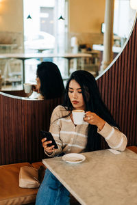 Young ethnic female with cup of hot drink text messaging on cellphone at cafeteria table against mirror