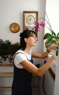 Side view of attentive female in eyeglasses checking leaves of potted plants growing on windowsill at home