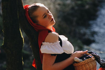 Young woman holding camera while standing against tree