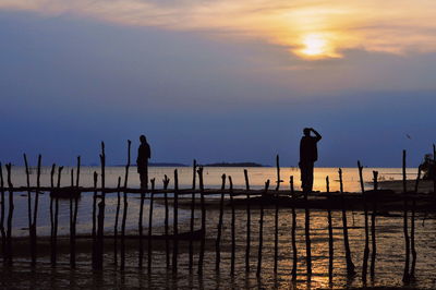 Silhouette men standing on wooden post by sea against sky during sunset