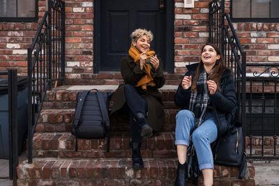 Happy female friends talking while sitting on steps in city during winter