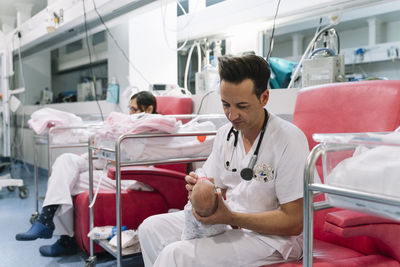 Doctors sitting on armchairs and feeding newborns from bottles holding heads in room with equipment in hospital