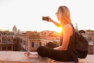 Side view of woman photographing in city against sky during sunset