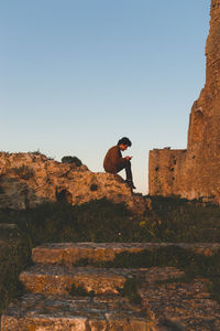 Man using mobile phone while sitting on rock during sunset