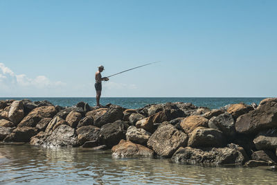 Man fishing on rock by sea against sky