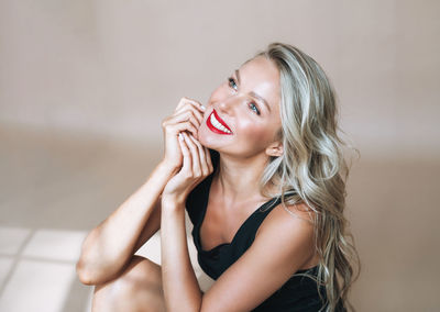 Portrait of happy blonde young woman with bright makeup in evening dress on beige background