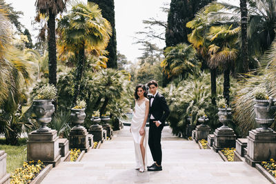 Happy lovers the bride and groom in wedding outfits walk among plants and palm trees in the old park