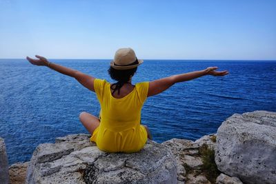 Rear view of woman sitting with arms outstretched on rock by sea against sky