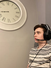 Boy wearing headphones sitting by clock on wall