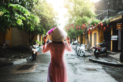 Rear view of woman wearing asian style conical hat while walking on wet street in city