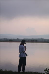 Rear view of man standing by lake against sky during sunset