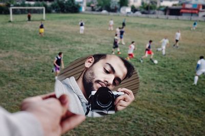 Reflection of man holding camera in mirror with boys playing soccer on field