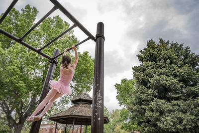 Rear view of girl in ballet costume hanging on monkey bars against cloudy sky at playground