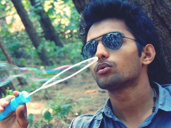 Close-up of young man wearing sunglasses blowing bubbles in forest