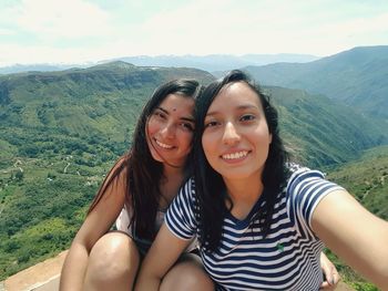 Portrait of smiling young female friends sitting on cliff against sky