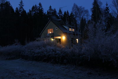 Illuminated house amidst trees and buildings against sky at night