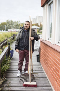 Portrait of man holding broom and equipment while standing by house