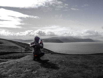 Rear view of man crouching while photographing on mobile phone over sea against sky