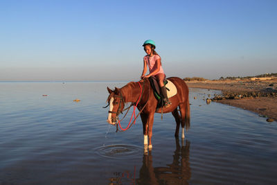 Girl riding horse in sea against blue sky