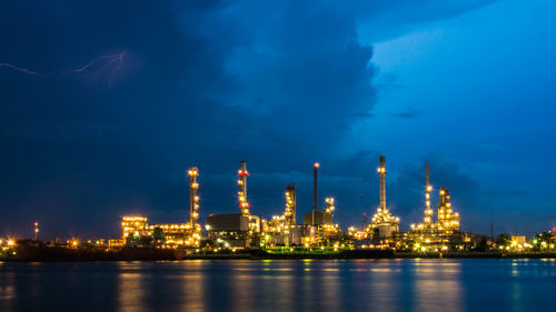 Illuminated factory by river against sky at night