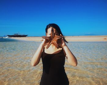 Woman covering face with dead starfish on shore against blue sky