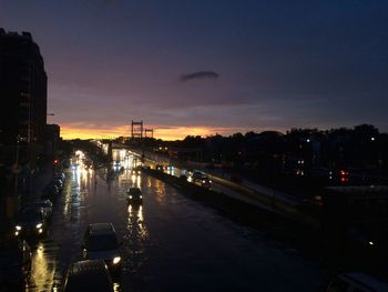 Illuminated city street by buildings against sky at sunset