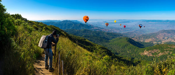Rear view of man photographing hot air balloons flying over landscape against sky