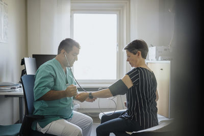 Male physician checking blood pressure of senior woman while sitting in clinic