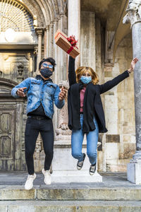 Young happy couple wearing mask in time of coronavirus jumping with a gift in hand