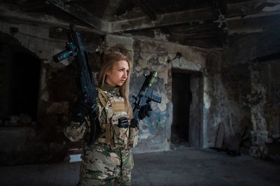 A woman in an army uniform aims to shoot a firearm in an abandoned building. 