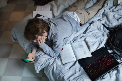 High angle view of teenage boy using mobile phone while lying by books and laptop on bed at home
