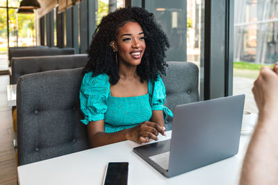 Cheerful young african american woman in turquoise top sitting at table with smartphone and working via netbook while chatting with cropped unrecognizable boyfriend in drinking coffee