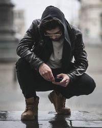 Man using mobile phone while crouching on wet footpath