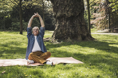 Smiling senior man with arms raised sitting on exercise mat at park