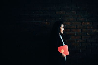 Side view of woman holding book while standing against brick wall