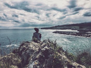 Woman sitting on rock formation while looking at sea against cloudy sky