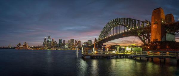 Illuminated bridge over river by buildings against sky at dusk