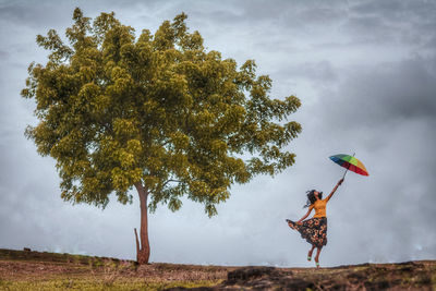 Full length of woman jumping with umbrella against cloudy sky