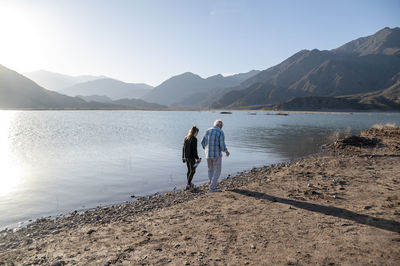 Senior man walking with his granddaughter along the lake shore with mountains.