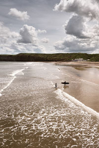 Two surfers walk out of the sea on to an empty beach under a dramatic sky.