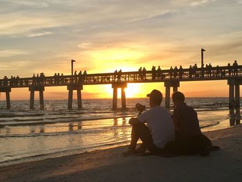 Rear view of men sitting on beach against sky during sunset