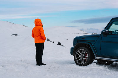 Man in winter orange jacket standing by car on snowy mountain during winter