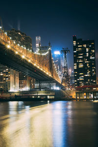 Illuminated bridge over river by buildings against sky at night