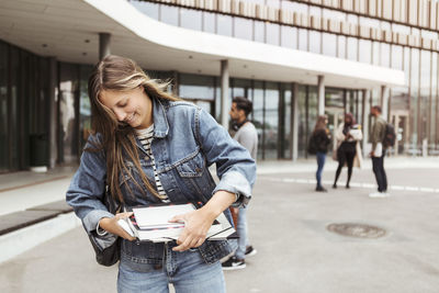 Smiling young woman holding books while standing in university campus