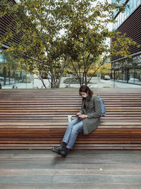 Full length of woman wearing mask sitting on bench outdoors