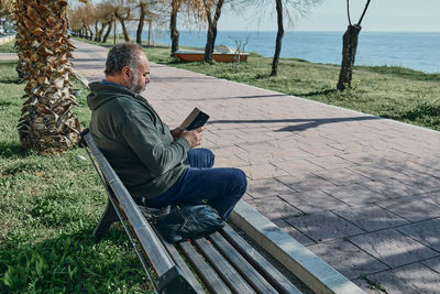 Mature man sitting alone on a park bench and using smartphone on a sunny day in a palm tree alley 